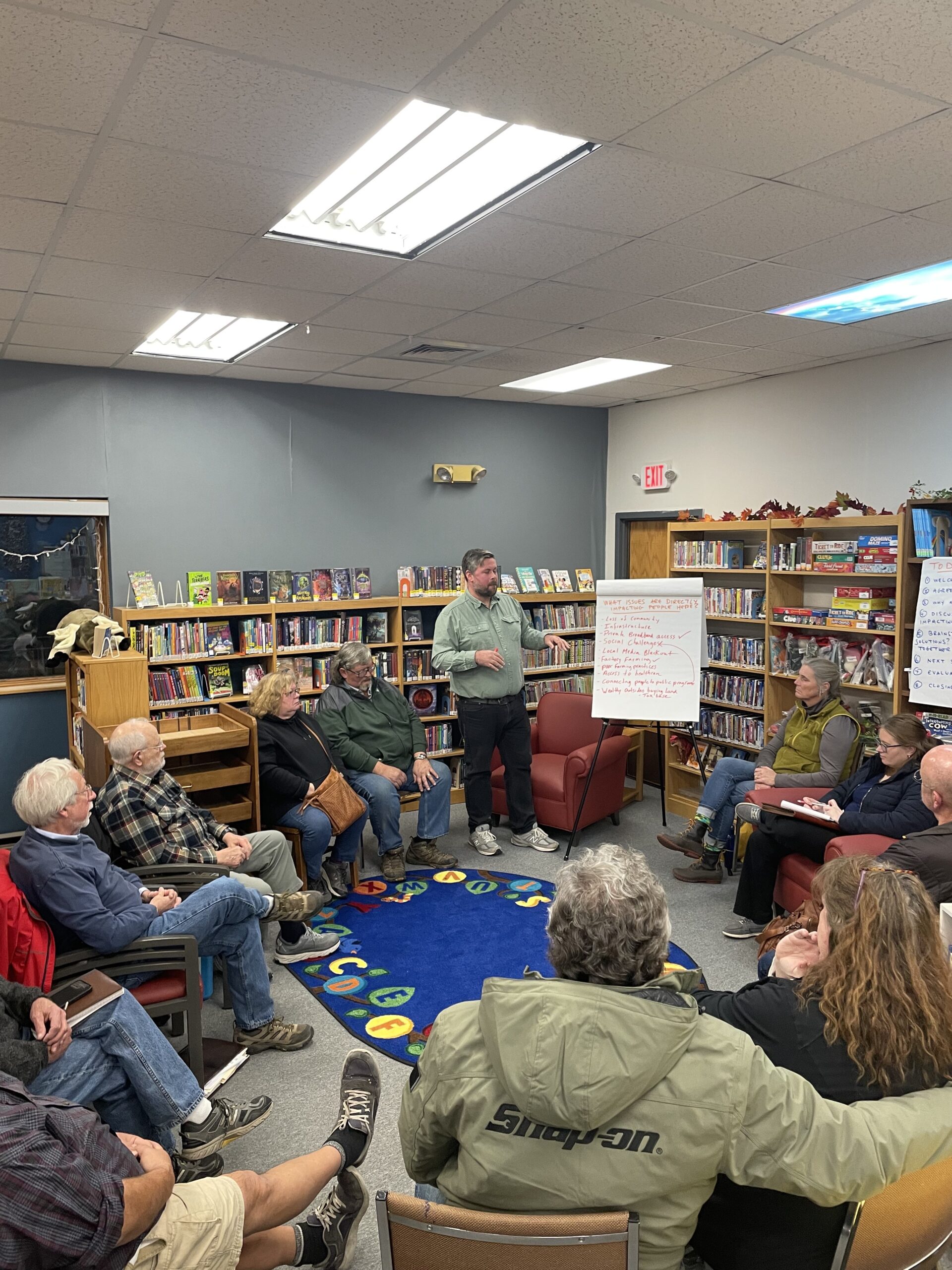 A group of people in jackets and flannels sit in a circle in a colorful corner of a small public library in Wisconsin
