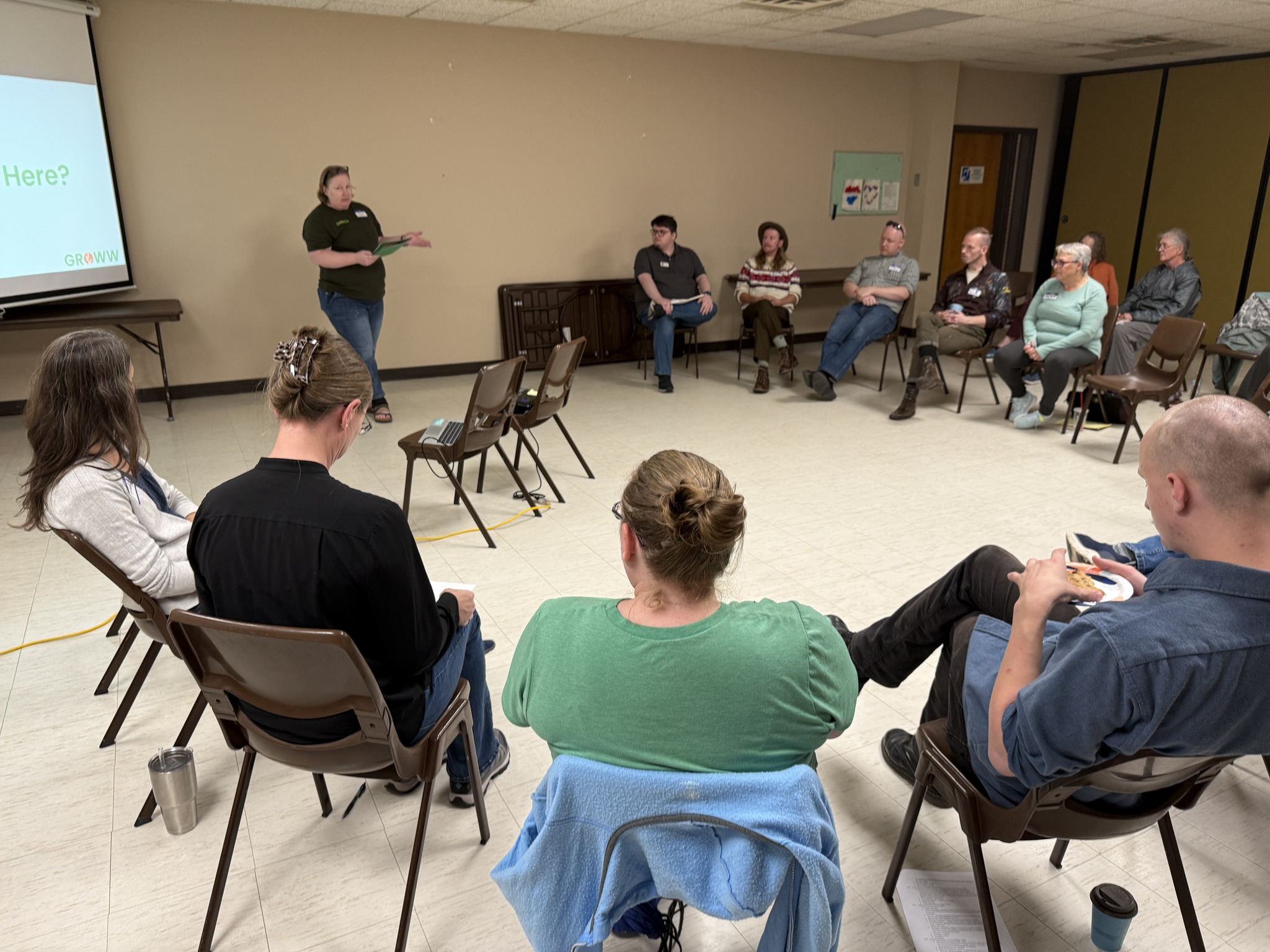 In a public meeting room, a group sits in a circle of folding chairs introducing themselves